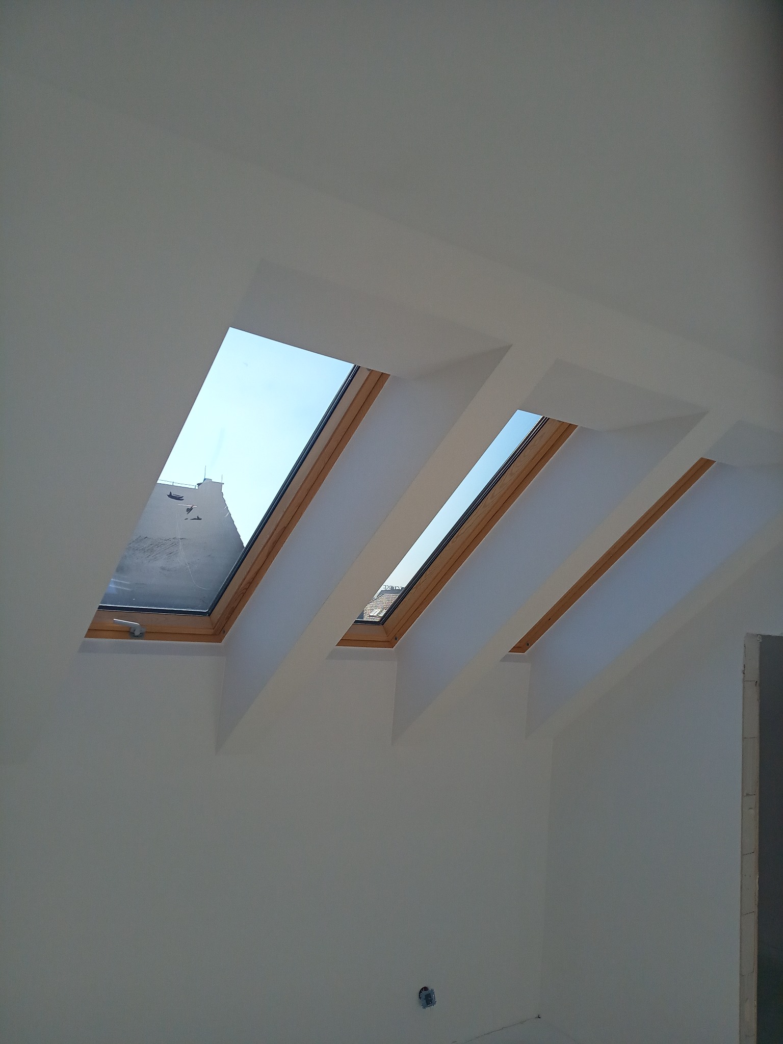 Dwa okna dachowe z drewnianymi ramami w białym, świeżo pomalowanym poddaszu, widok na budynek z kominem przez jedno z okien.