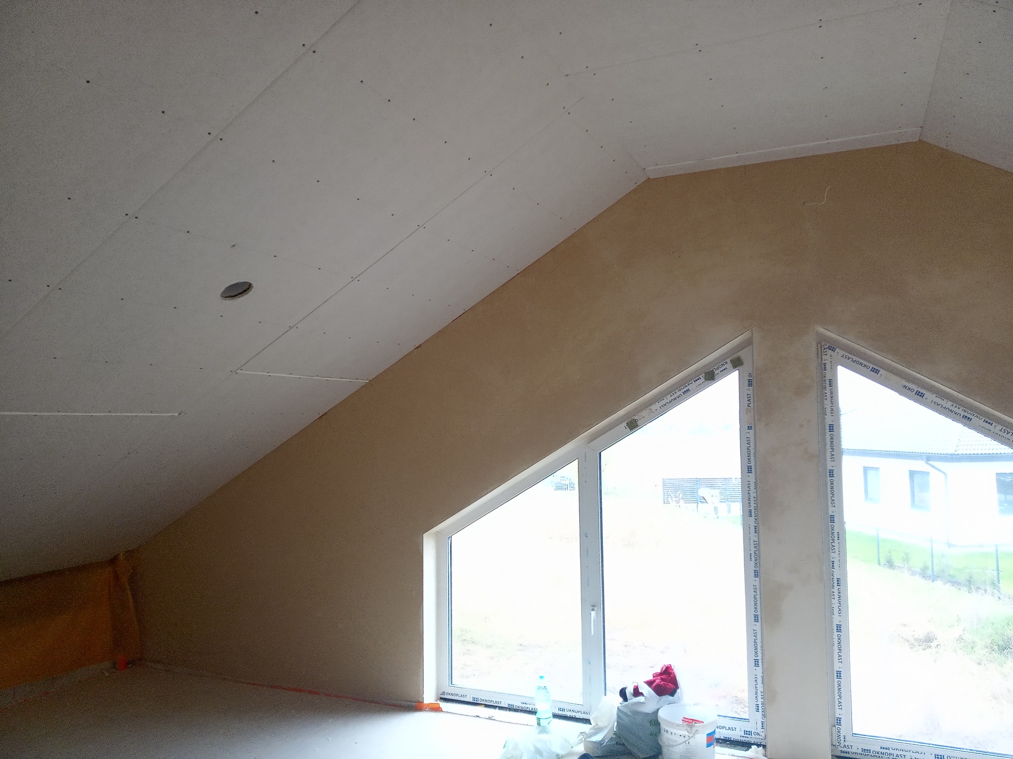 Poddasze z oknami trójkątnymi, ściany wykończone gładzią w kolorze beżowym, sufit z zamontowaną płytą kartonowo-gipsową, widoczne ślady po szpachlowaniu, na podłodze materiały budowlane i butelka...
