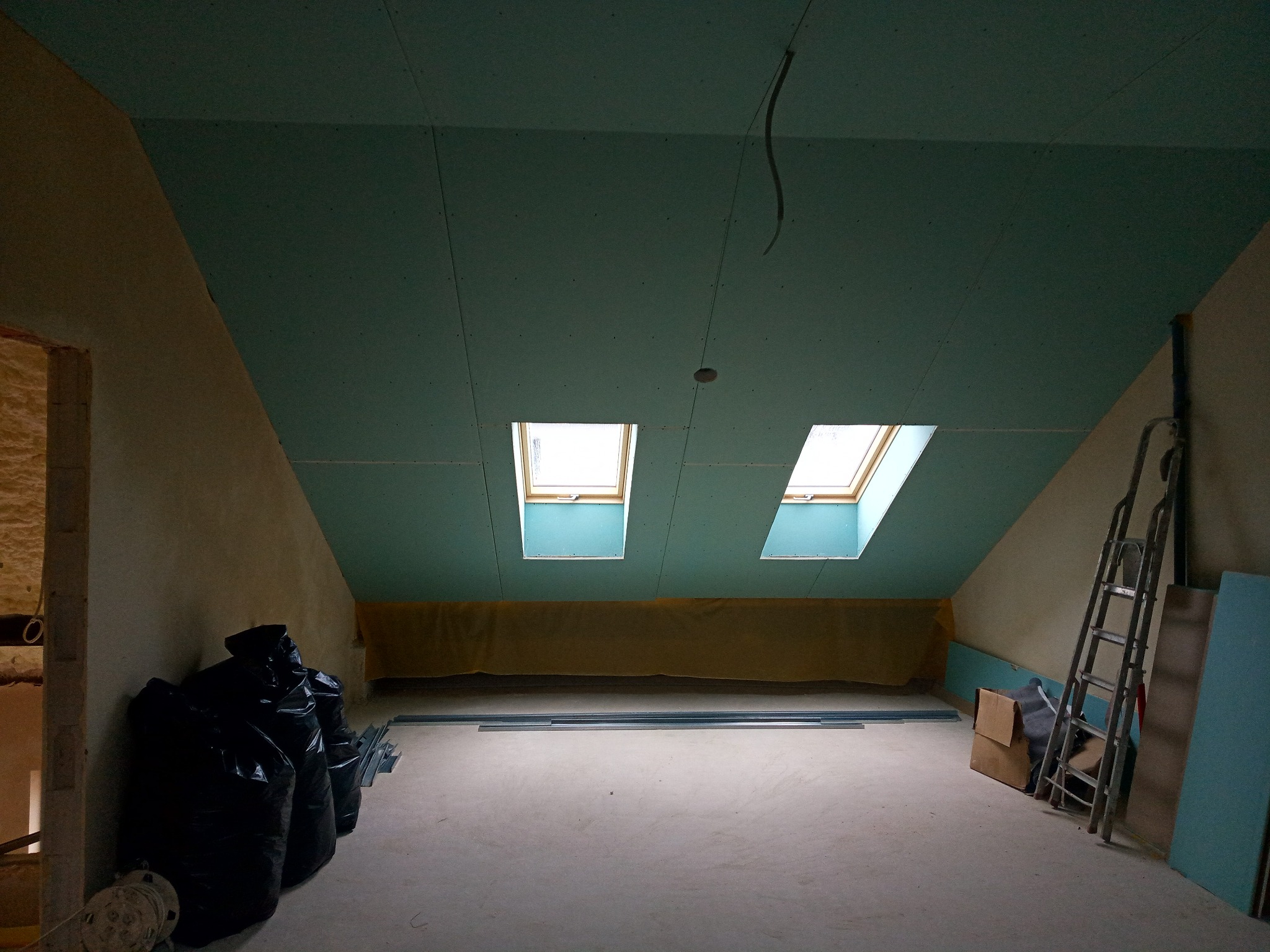 Poddasze w trakcie prac wykończeniowych z zamontowanymi oknami dachowymi, ściany częściowo wykończone, widoczne worki z odpadami, drabina i płyty.