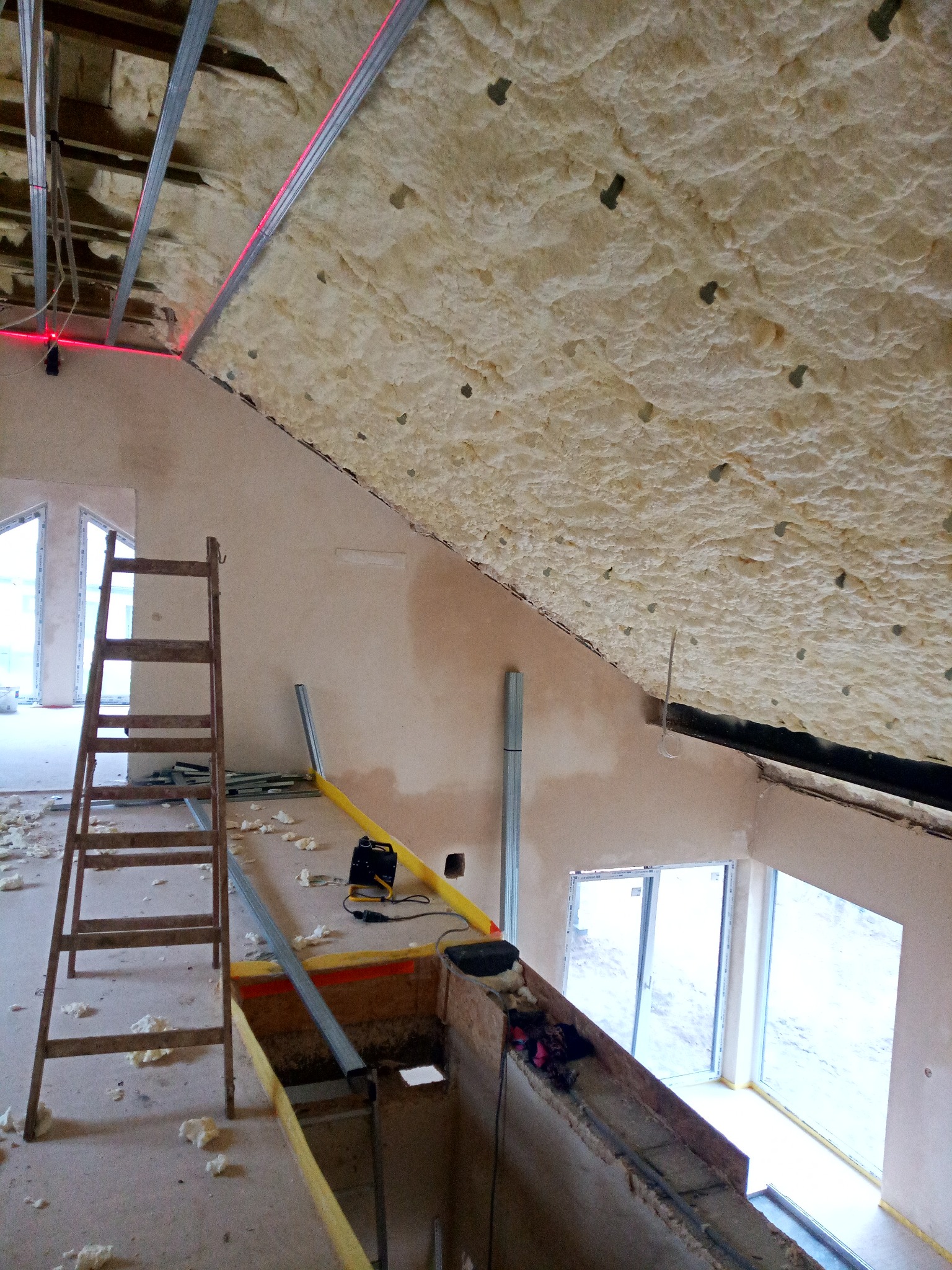 Poddasze w trakcie prac izolacyjnych, widoczna piana poliuretanowa na skośnym dachu, metalowy stelaż, drabina drewniana i poziomica laserowa wyznaczająca linię montażu.