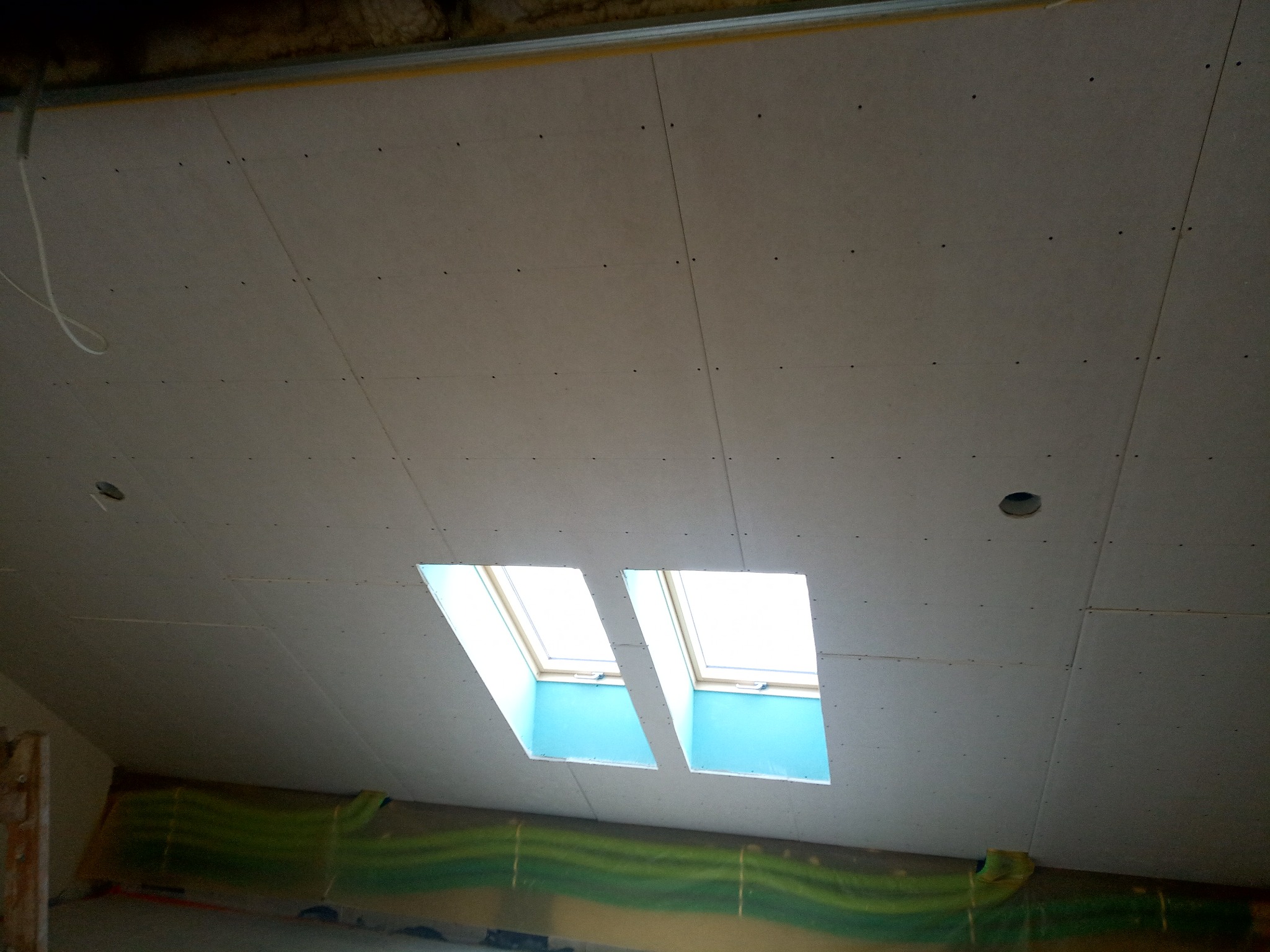 Poddasze z zamontowanymi płytami, widoczne dwa okna dachowe i otwory na oświetlenie, zabezpieczona folią podłoga.