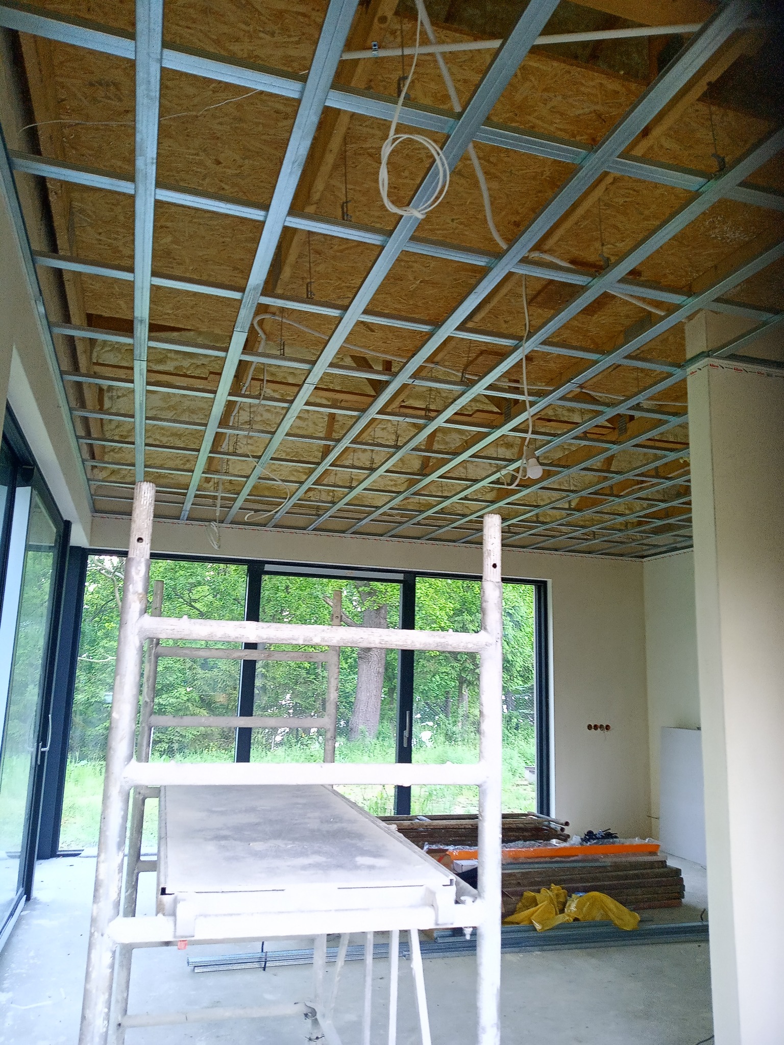 Sufit w trakcie montażu: widoczne profile metalowe tworzące szkielet konstrukcji, przestrzeń wypełniona materiałem izolacyjnym, drabina rusztowaniowa w pomieszczeniu z dużymi oknami.