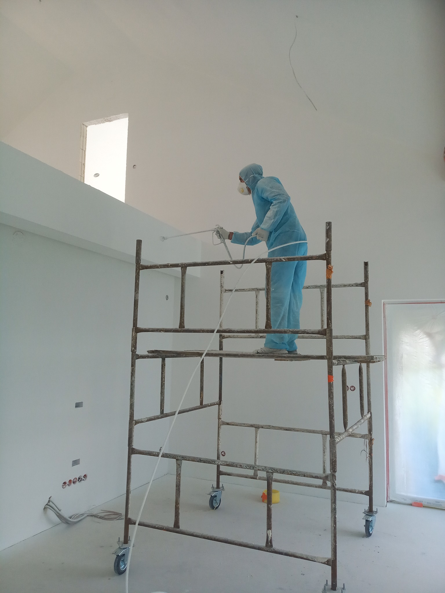 Pracownik w kombinezonie ochronnym maluje sufit pistoletem natryskowym stojąc na rusztowaniu w jasnym, wysokim pomieszczeniu z oknem w górnej części ściany.