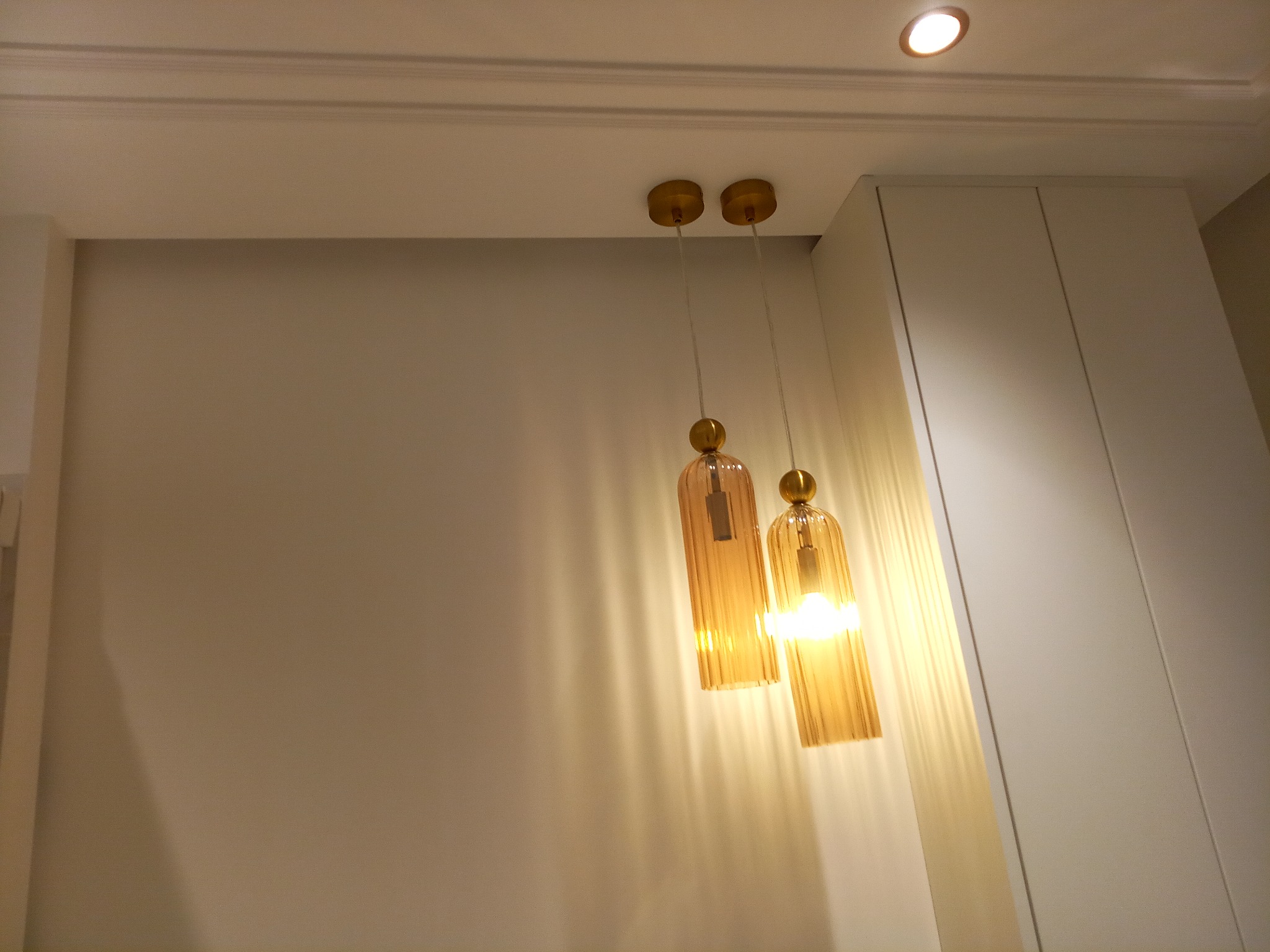 Dwa wiszące szklane klosze w odcieniach bursztynu z żarówkami, złote mocowania, widok na biały sufit z listwą dekoracyjną i wbudowanym oświetleniem oraz fragment białej szafy.