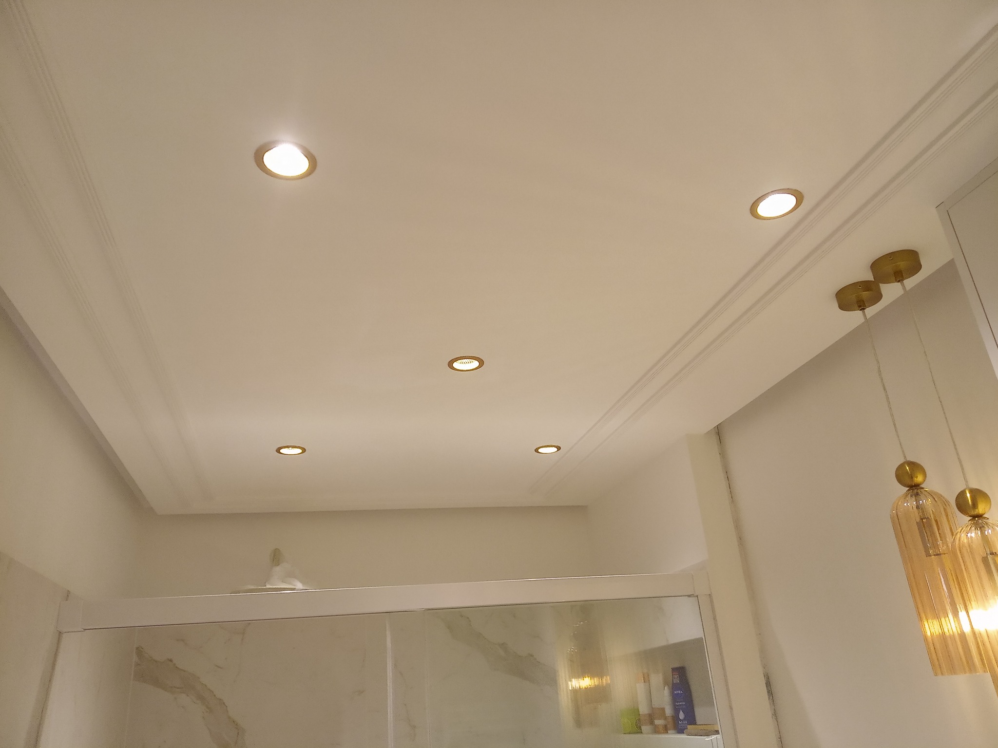 Sufit w łazience z oświetleniem punktowym i dekoracyjnymi listwami, widoczny fragment kabiny prysznicowej z marmurowym wzorem oraz ozdobne lampy wiszące.