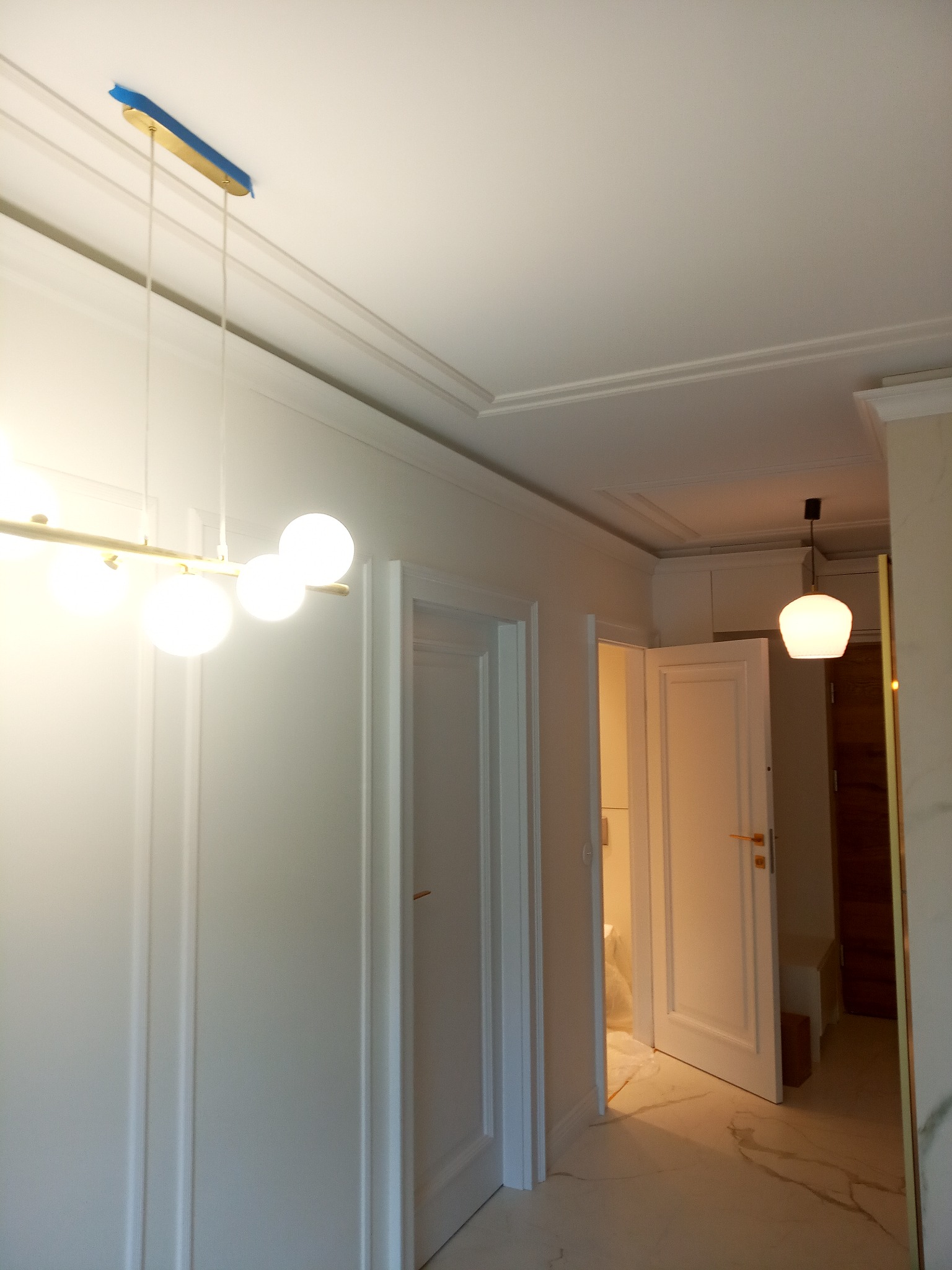 Wnętrze po remoncie z otwartymi białymi drzwiami, widoczne listwy przysufitowe, nowoczesne lampy wiszące i marmurowe płytki podłogowe.