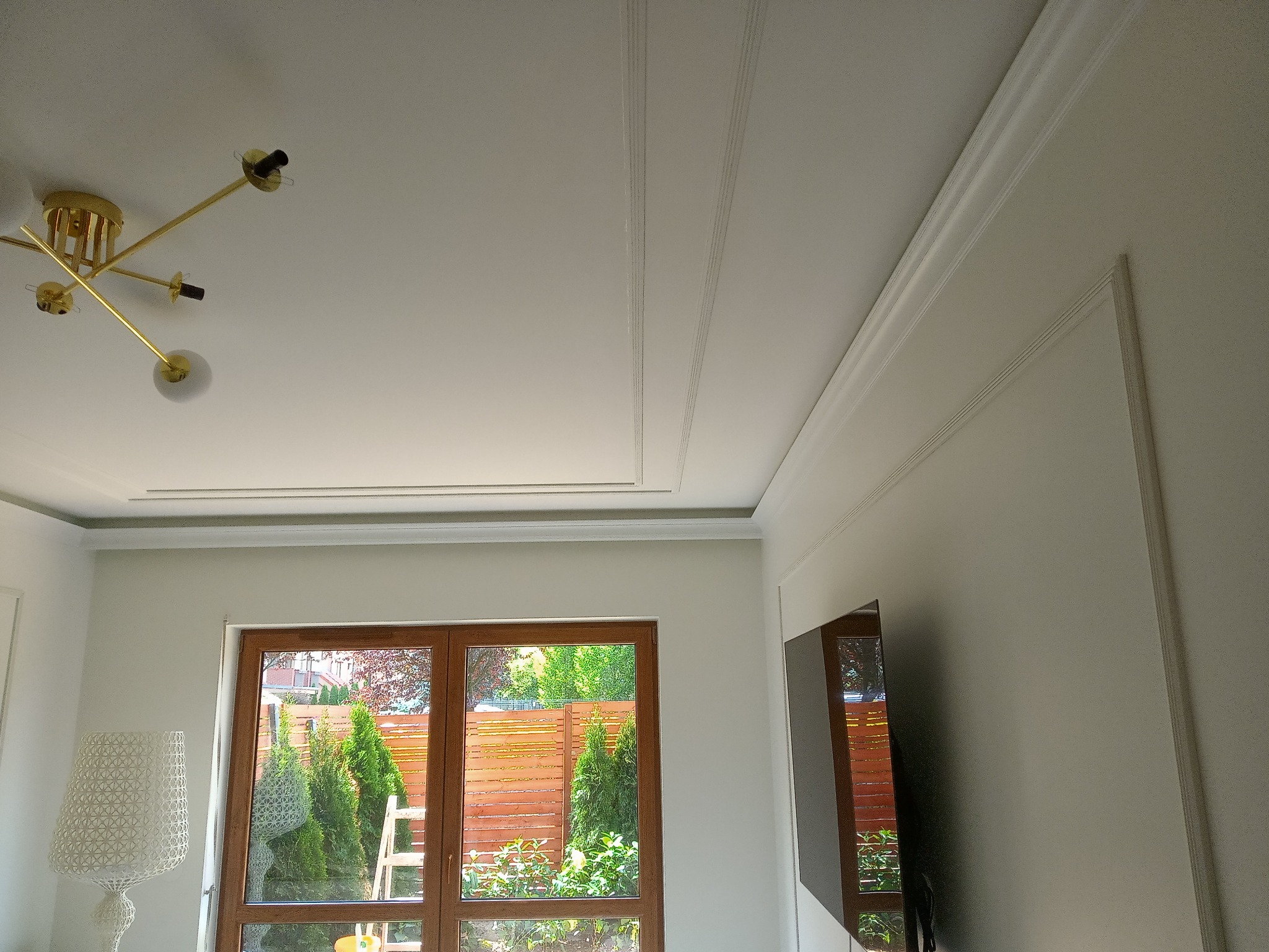 Nowoczesne wnętrze z białym sufitem z dekoracyjnymi listwami, złotą lampą sufitową i dużym oknem z widokiem na zielony ogród.