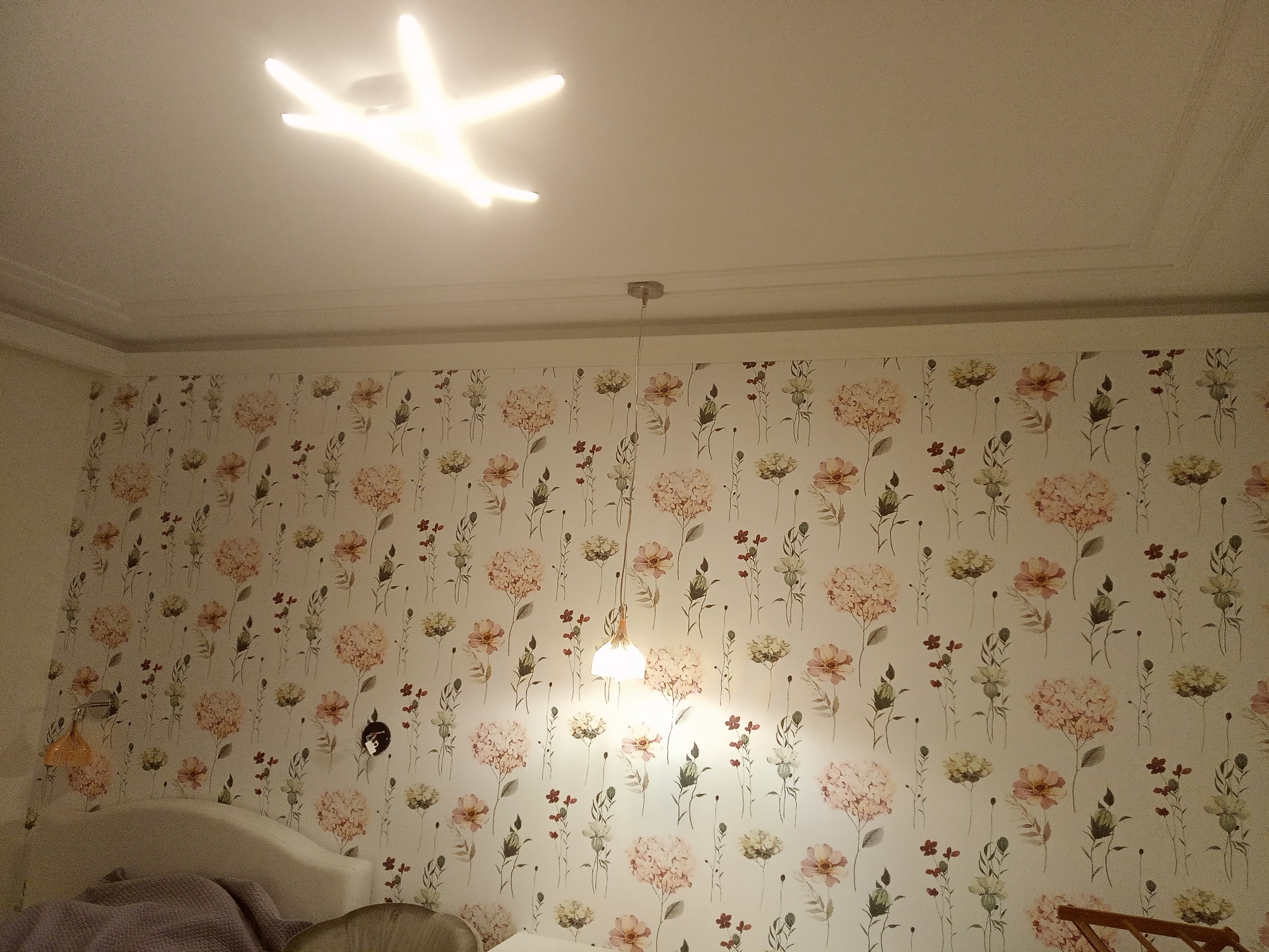 Kwiatowa tapeta w pokoju we Wrocławiu z nowoczesnym oświetleniem sufitowym w kształcie gwiazdy i wiszącą lampą.