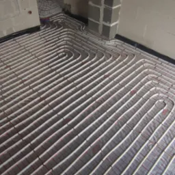 Białe rury ogrzewania podłogowego ułożone w pętle na folii z siatką, widoczne fragmenty ścian i sufitu w surowym stanie.