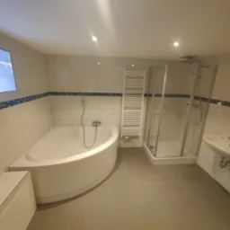 Jasna łazienka z narożną wanną, kabiną prysznicową i grzejnikiem drabinkowym, wykończona białymi płytkami z niebieskim akcentem dekoracyjnym.