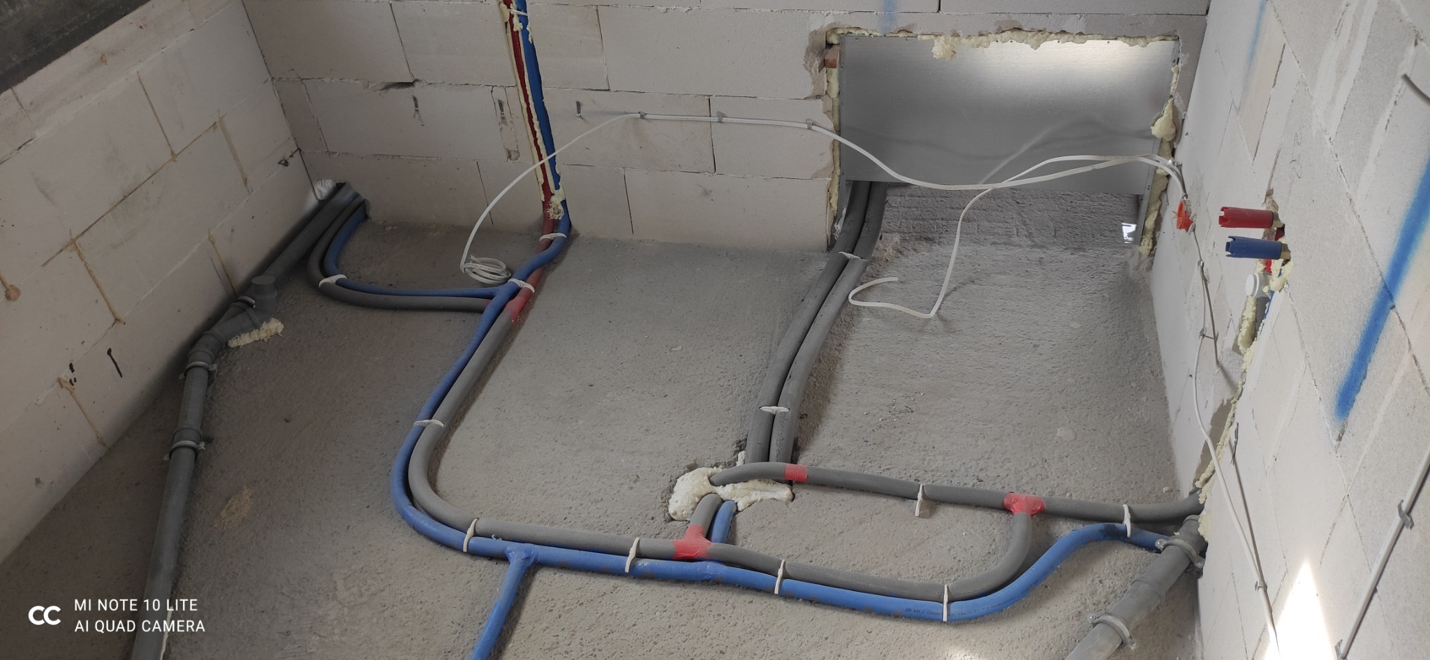 Surowe wnętrze z instalacją wodno-kanalizacyjną w trakcie montażu: niebieskie i szare rury prowadzone po betonowej podłodze i ścianach z białego bloczka, zabezpieczone pianką montażową w miejscach...