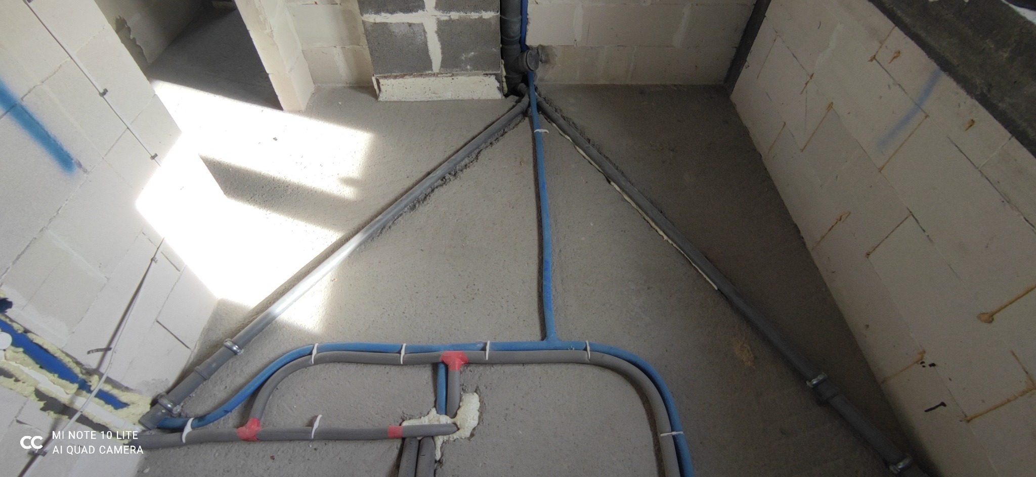 Szare i niebieskie rury instalacyjne ułożone w bruzdach w wylewce podłogowej, ściany z betonu komórkowego, widoczne piankowanie.