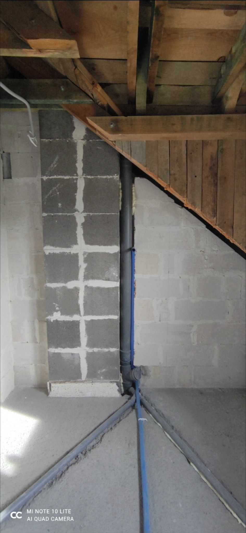 Szara rura kanalizacyjna pionowo przy ścianie z bloczków betonowych, poziome rury kanalizacyjne na podłodze, surowy stan wykończenia wnętrza, drewniana konstrukcja dachu