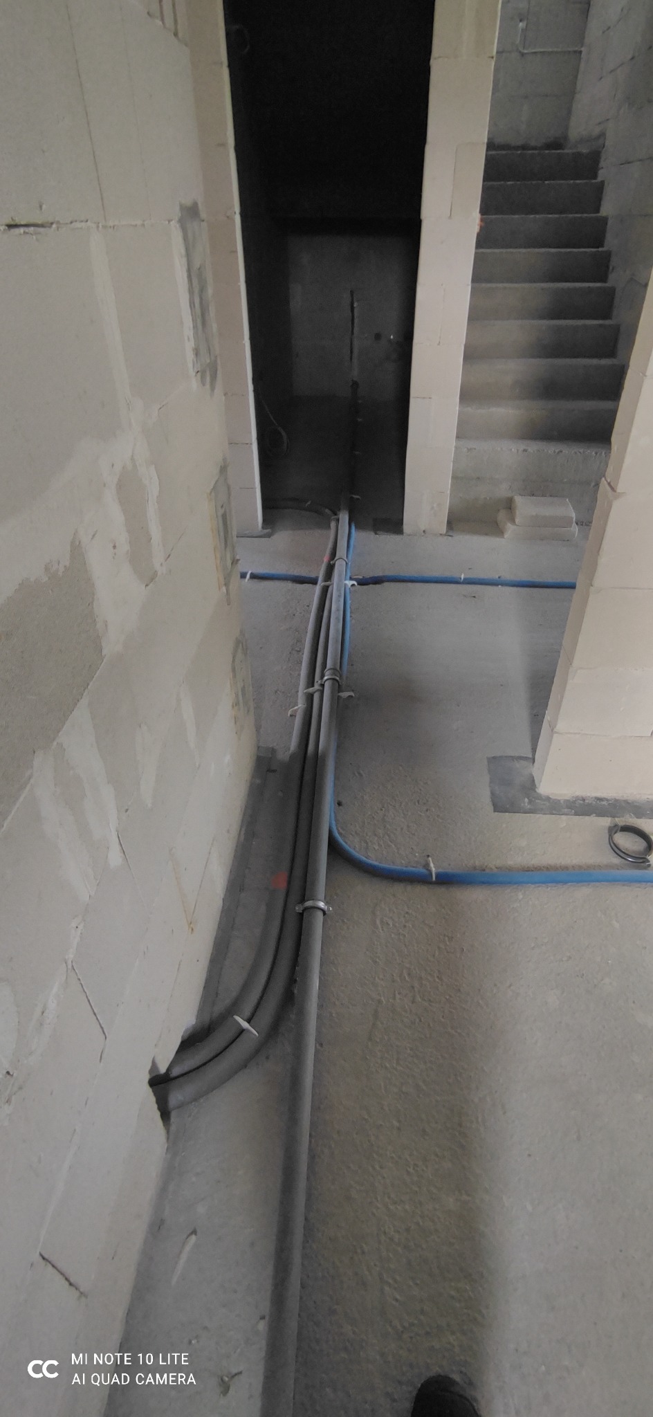 Szare rury instalacyjne prowadzone wzdłuż ściany z betonu komórkowego w surowym stanie, niebieska rura poprowadzona po podłodze, widoczne schody w tle, wnętrze w stanie deweloperskim.