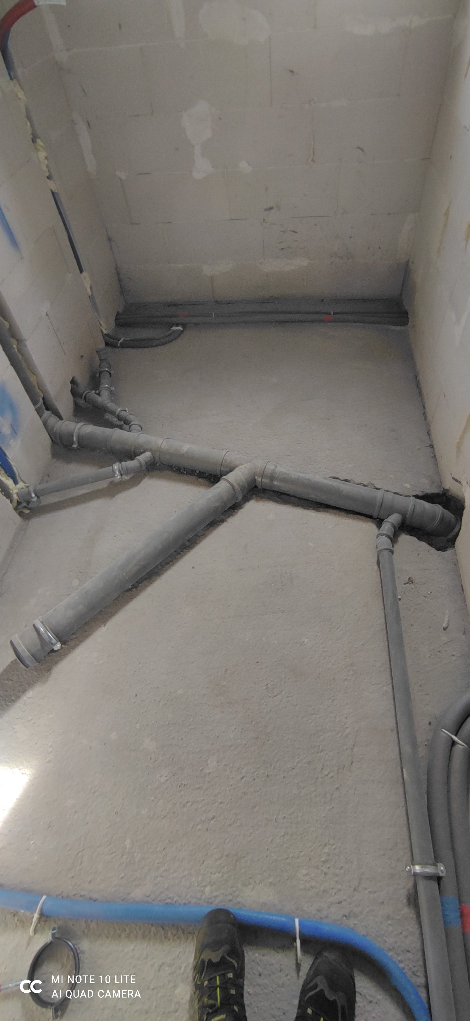 Szare rury kanalizacyjne z metalowymi obejmami, ułożone na surowym betonowym podłożu w trakcie instalacji, widoczne fragmenty nieotynkowanych ścian z betonu komórkowego i fragment butów na dole kadru.