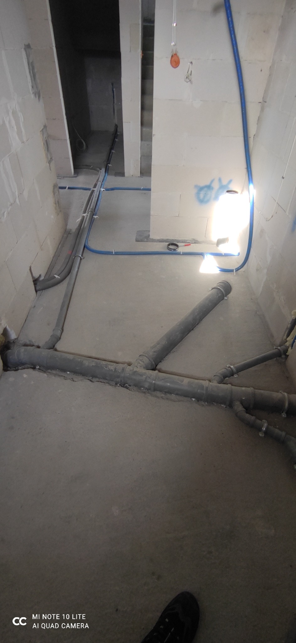 Szare rury kanalizacyjne z licznymi rozgałęzieniami ułożone na surowym betonowym podłożu w trakcie prac instalacyjnych w pomieszczeniu z bloczków betonowych. Widoczne także niebieskie rury...