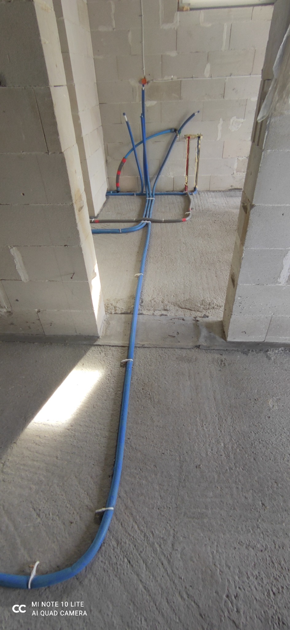 Niebieskie rury instalacji wodnej i grzewczej w surowym stanie podłogi i ścian z betonu komórkowego, widoczne złącza i mocowania do ściany.