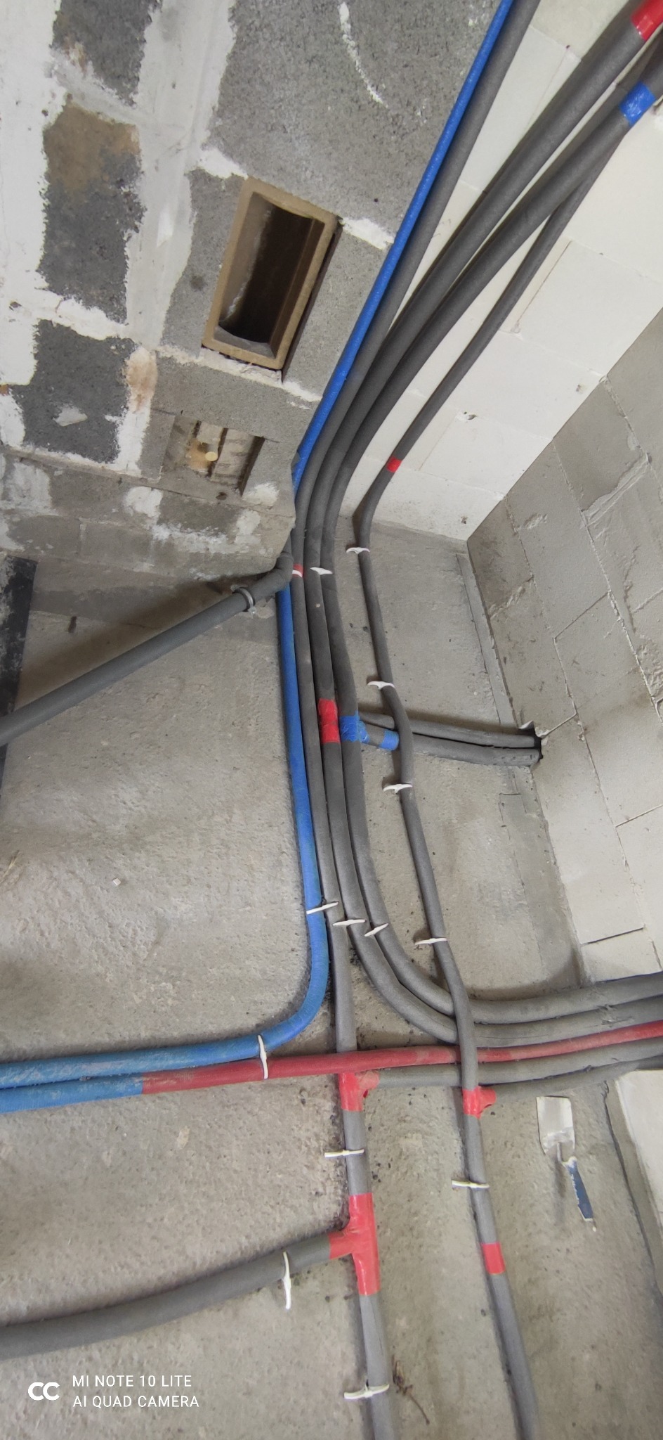 Szare i kolorowe rury instalacyjne, niektóre z izolacją, prowadzone wzdłuż surowych ścian z betonu komórkowego i sufitu w trakcie montażu.