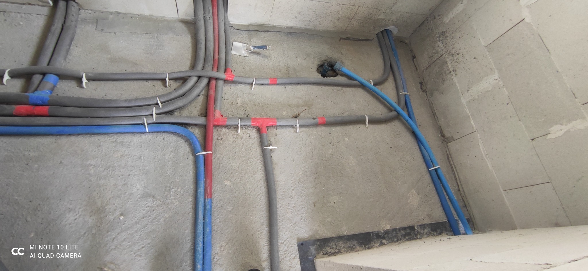 Szare, zaizolowane rury instalacji grzewczej i niebieskie rury wodne przymocowane do surowej, betonowej ściany za pomocą białych opasek zaciskowych, widoczna szpachelka w tynku.