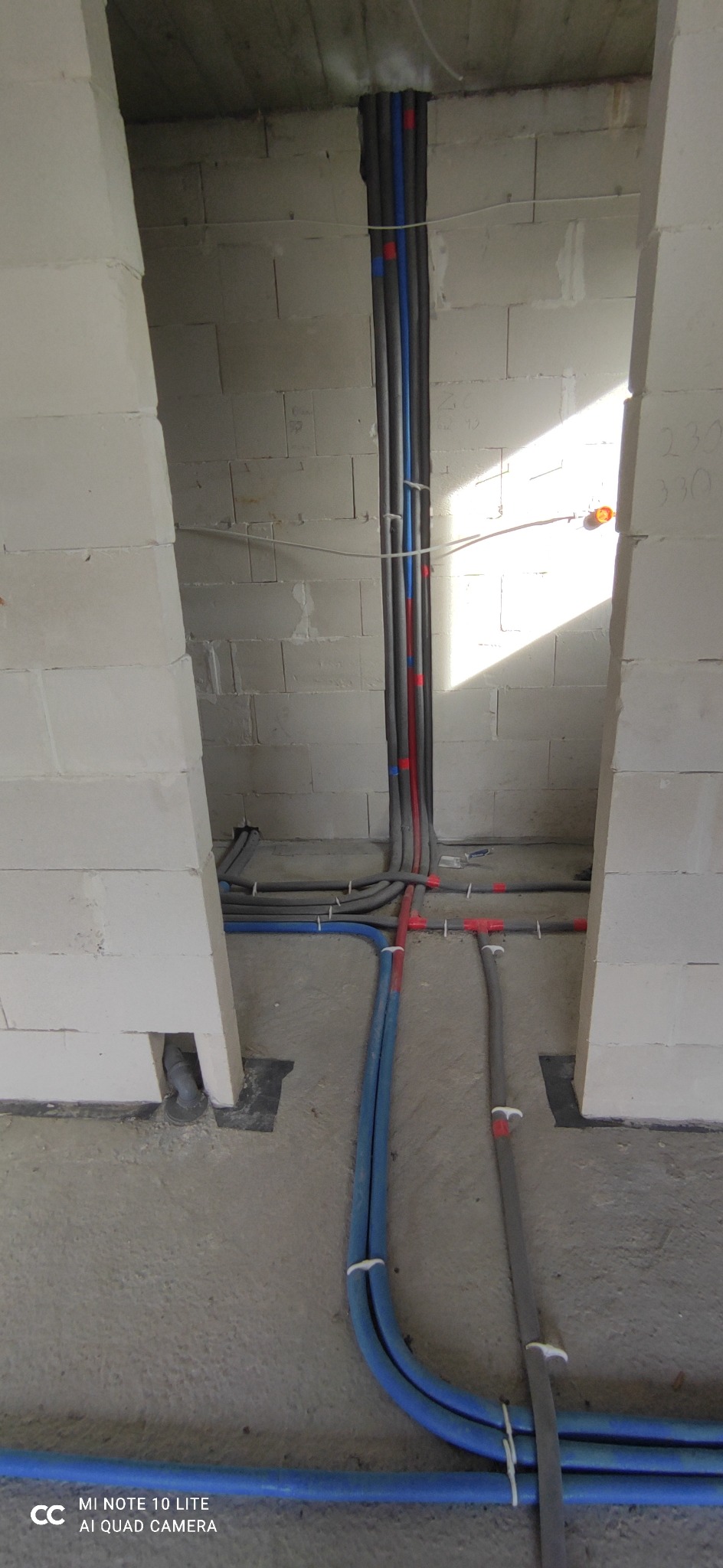 Surowe wnętrze z instalacją wodną i grzewczą w ścianach z betonu komórkowego; widoczne rury w izolacji, niebieskie rury wodne oraz mocowania.