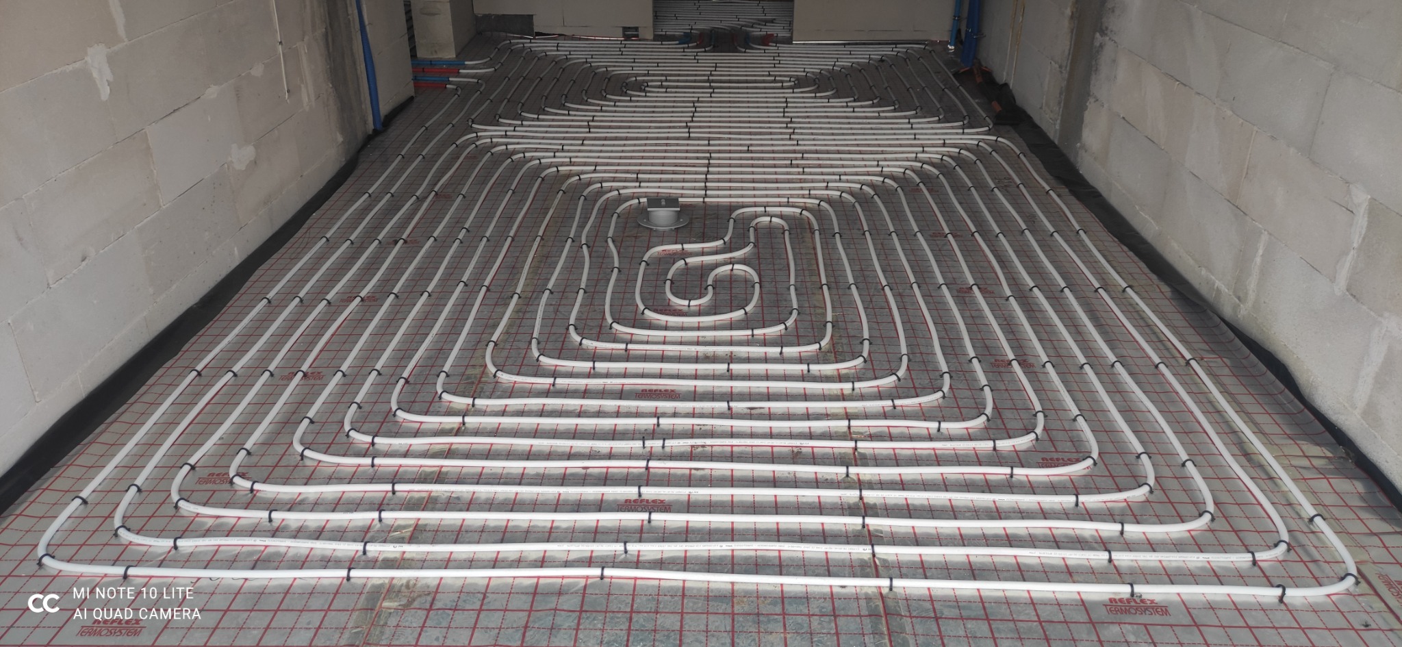 Białe rury ogrzewania podłogowego ułożone spiralnie na izolacji z siatką w pomieszczeniu z surowymi ścianami z betonu komórkowego.