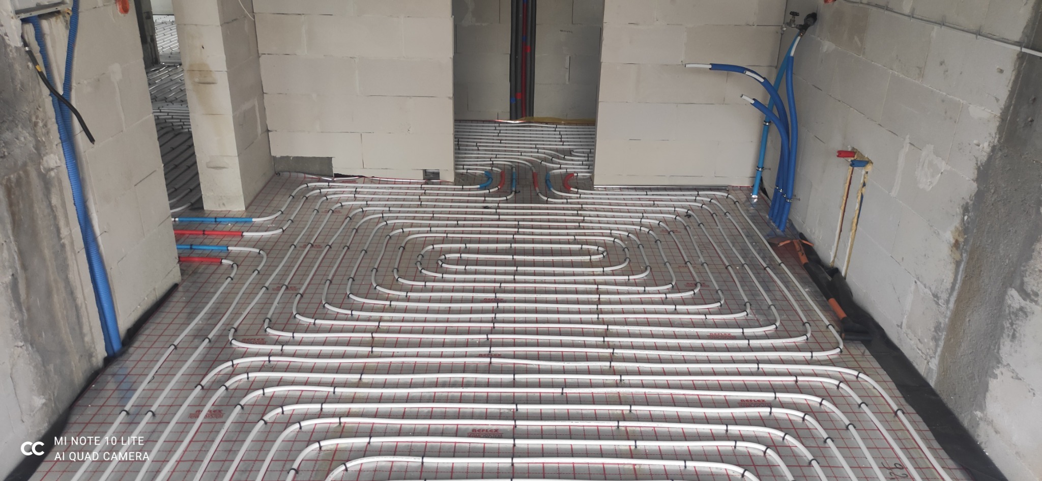 Rozłożone rury ogrzewania podłogowego na siatce w surowym wnętrzu, widoczne nieotynkowane ściany i wystające rury instalacji wodnej.
