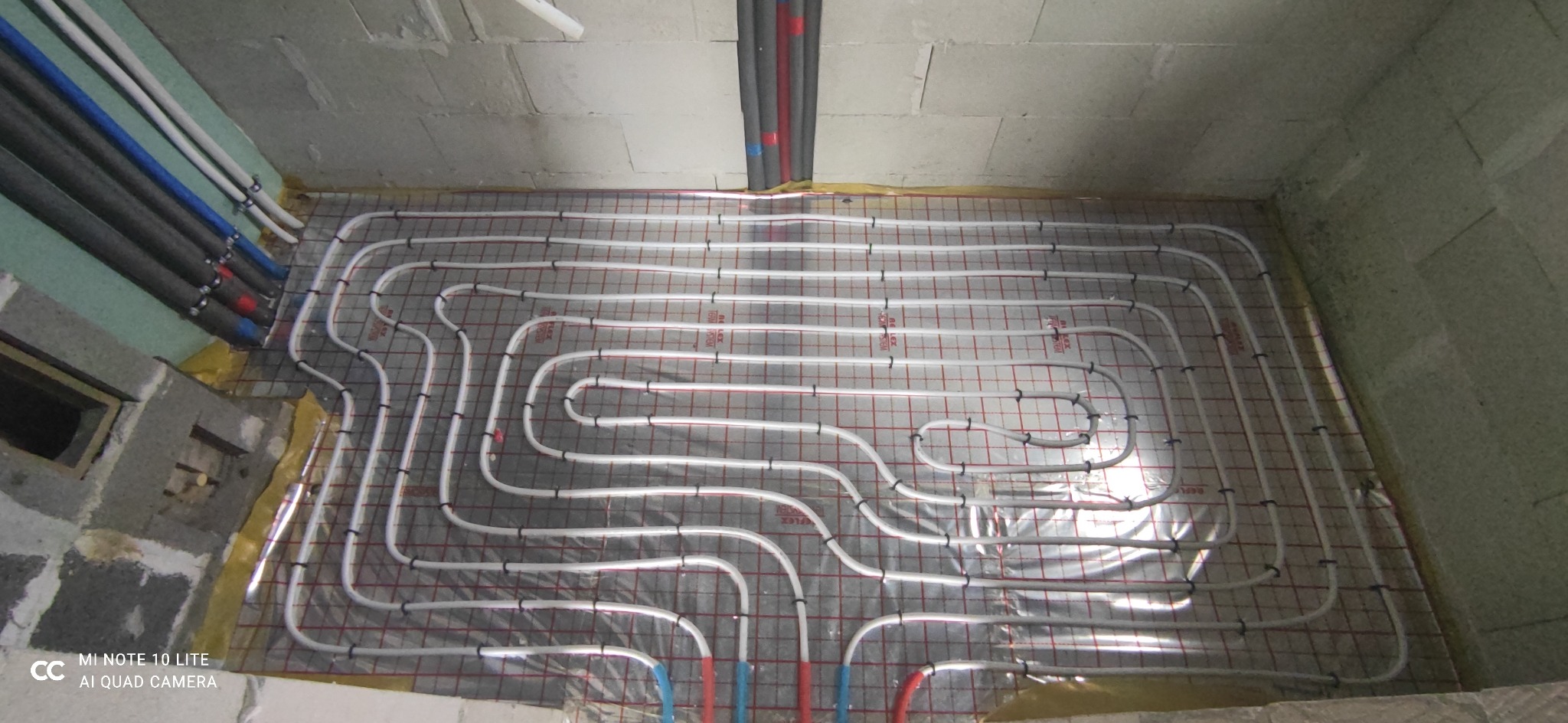 Białe rury ogrzewania podłogowego ułożone na folii z czerwoną siatką, widoczne szare ściany w surowym stanie i rury instalacyjne w izolacji przy ścianie.