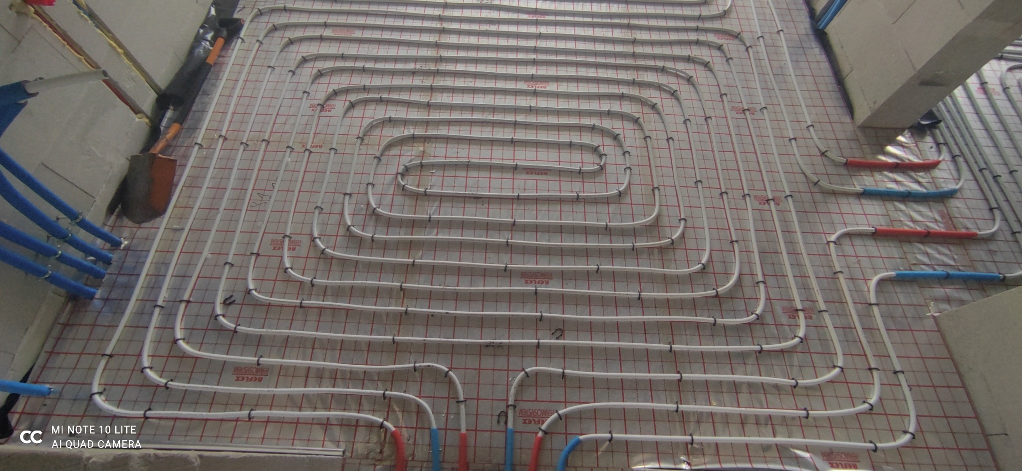 Rozłożone białe rurki ogrzewania podłogowego na folii z czerwoną kratką, widoczne niebieskie rury doprowadzające i narzędzie przy ścianie.