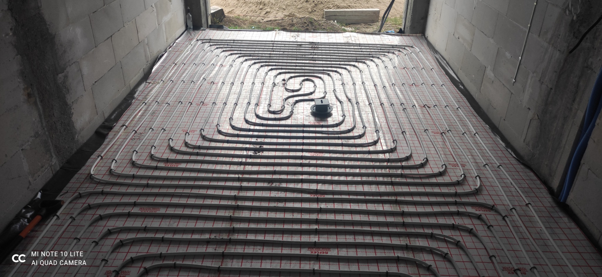 Rozłożone rury ogrzewania podłogowego na izolacji z siatką w surowym wnętrzu, widoczne przyłącze do rozdzielacza, ściany z betonu komórkowego.