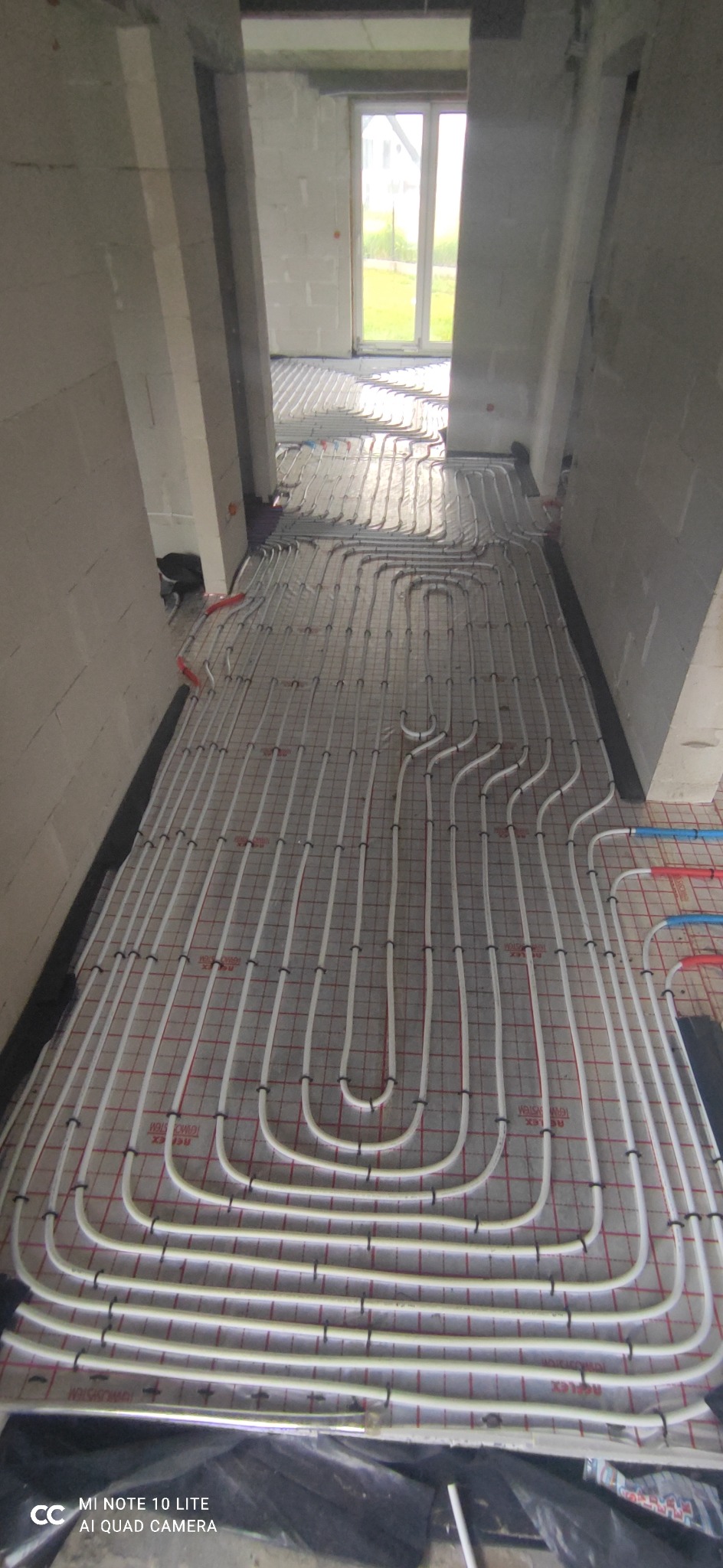Białe rury ogrzewania podłogowego ułożone na izolacji z siatką w surowym wnętrzu budynku w trakcie budowy, widoczne okno z zielenią na zewnątrz.