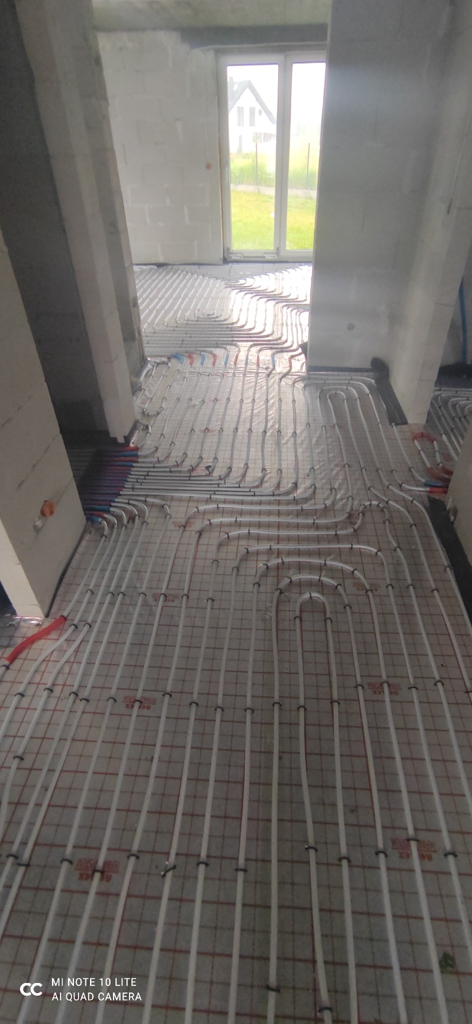 Białe rurki ogrzewania podłogowego ułożone na folii z siatką w surowym wnętrzu budynku w trakcie budowy, widok na okno i zieleń na zewnątrz.