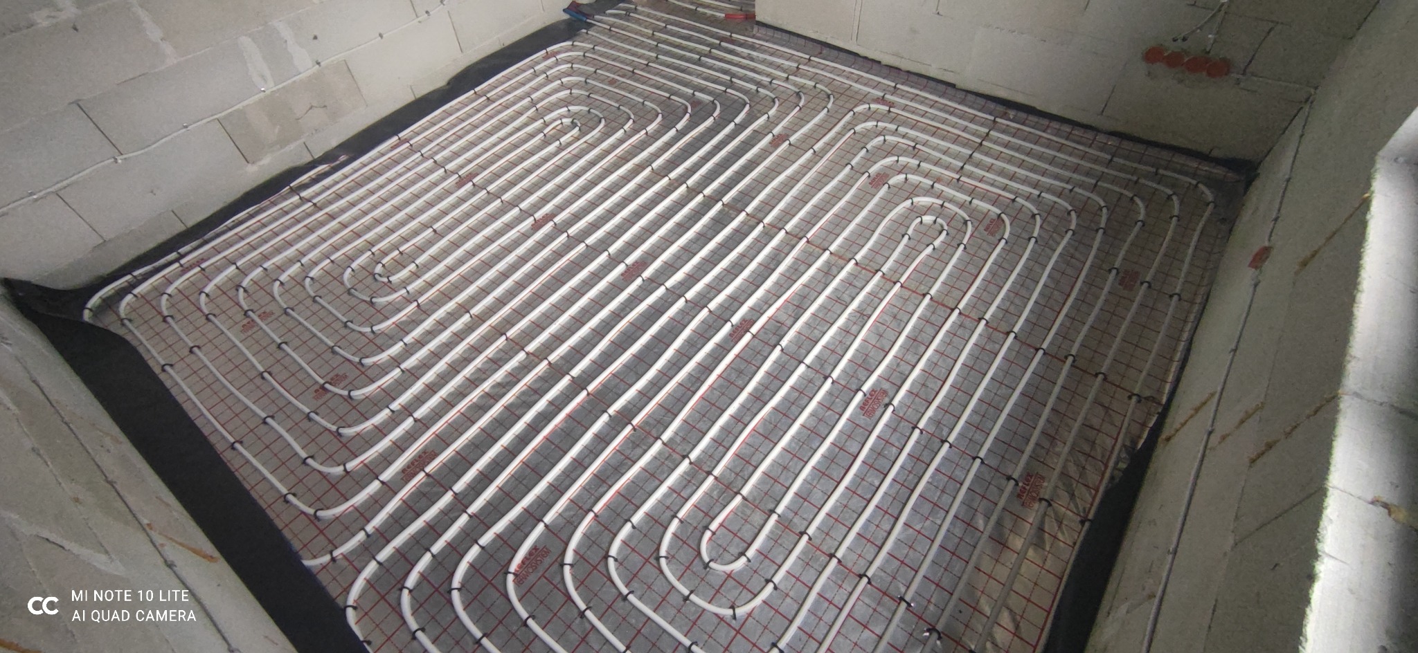 Białe rury ogrzewania podłogowego ułożone spiralnie na szarej macie z czerwoną siatką w surowym pomieszczeniu z betonowymi ścianami.