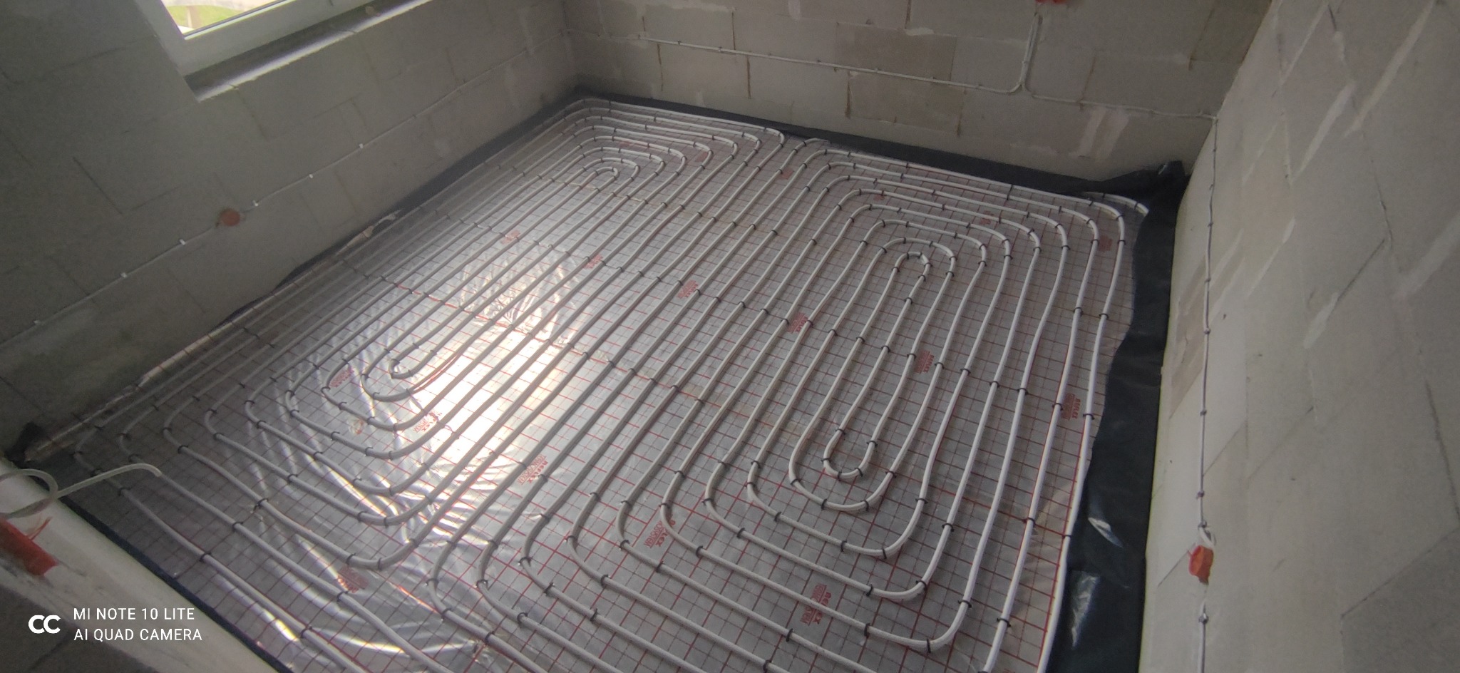 Białe rury ogrzewania podłogowego ułożone w pętle na folii z siatką w surowym pomieszczeniu z betonowymi ścianami i oknem u góry.