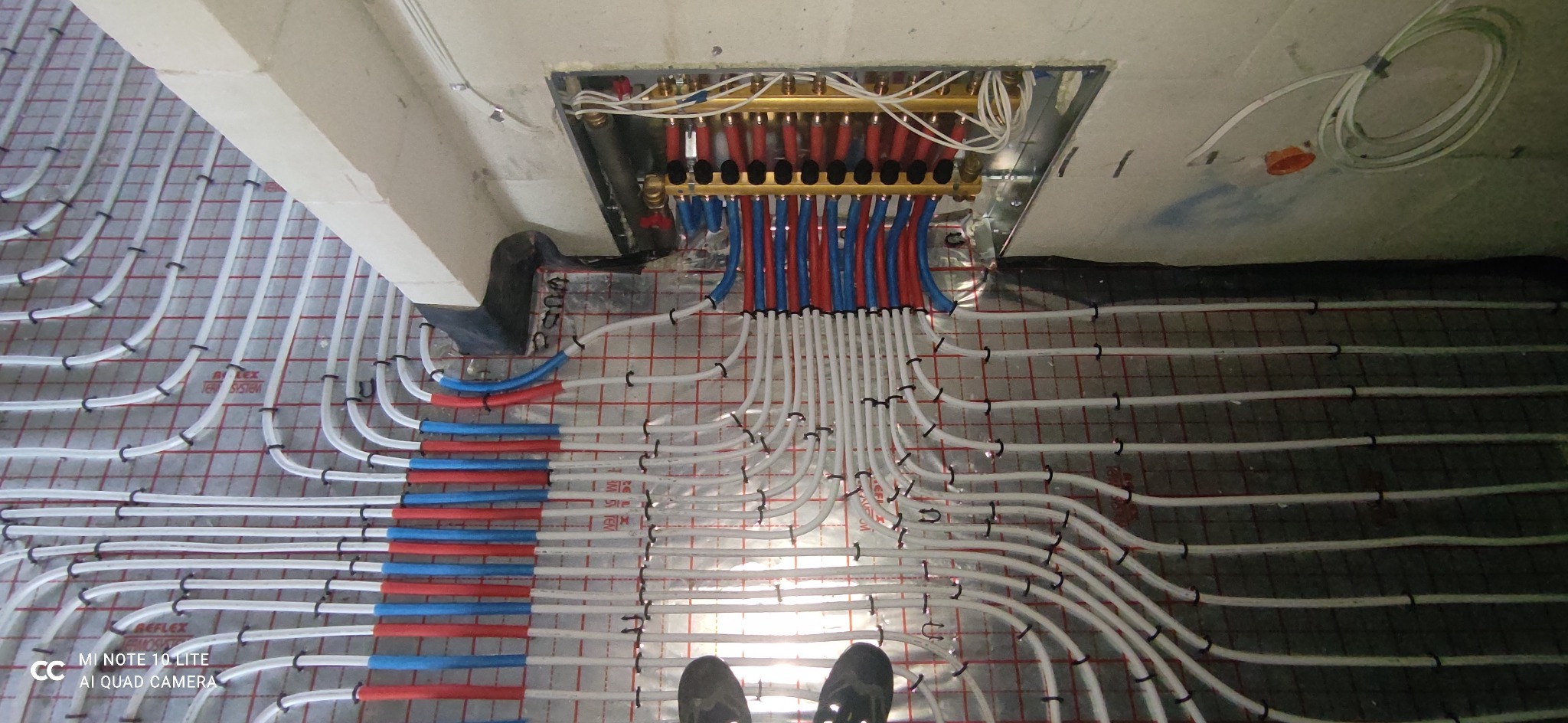 Rozdzielacz ogrzewania podłogowego z rurami rozprowadzonymi na folii z rastrem, widok z góry na etapie montażu, buty robocze w dolnej części kadru.