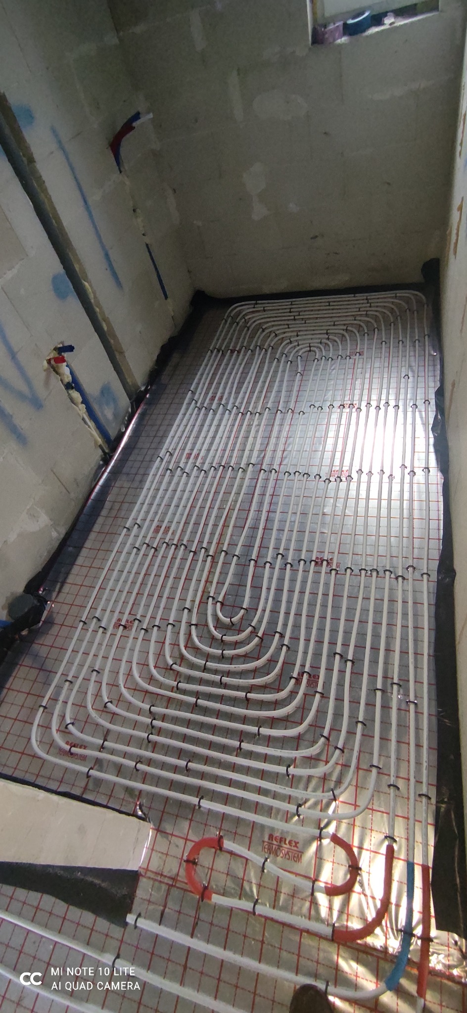 Białe rurki ogrzewania podłogowego ułożone spiralnie na folii z siatką, widoczne fragmenty ścian i instalacji w surowym stanie.