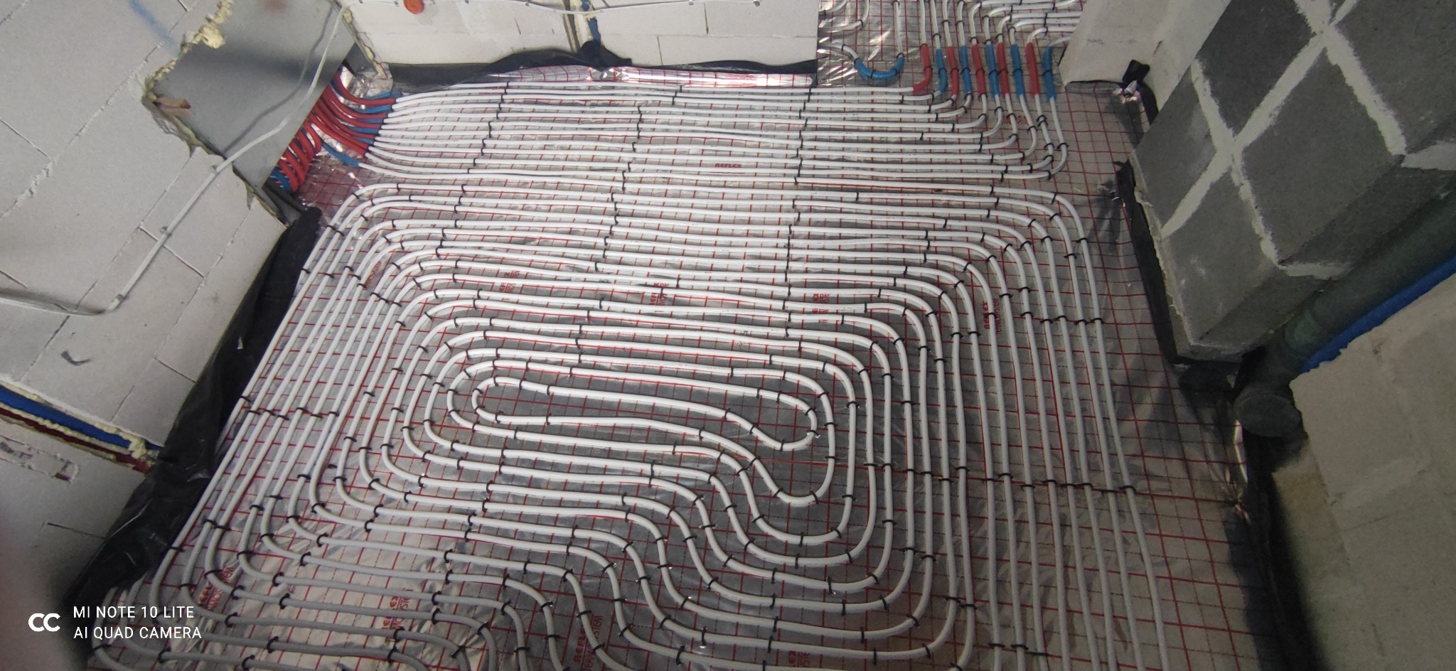 Białe rury ogrzewania podłogowego ułożone spiralnie na folii refleksyjnej z czerwoną siatką, widoczne fragmenty ścian w surowym stanie.