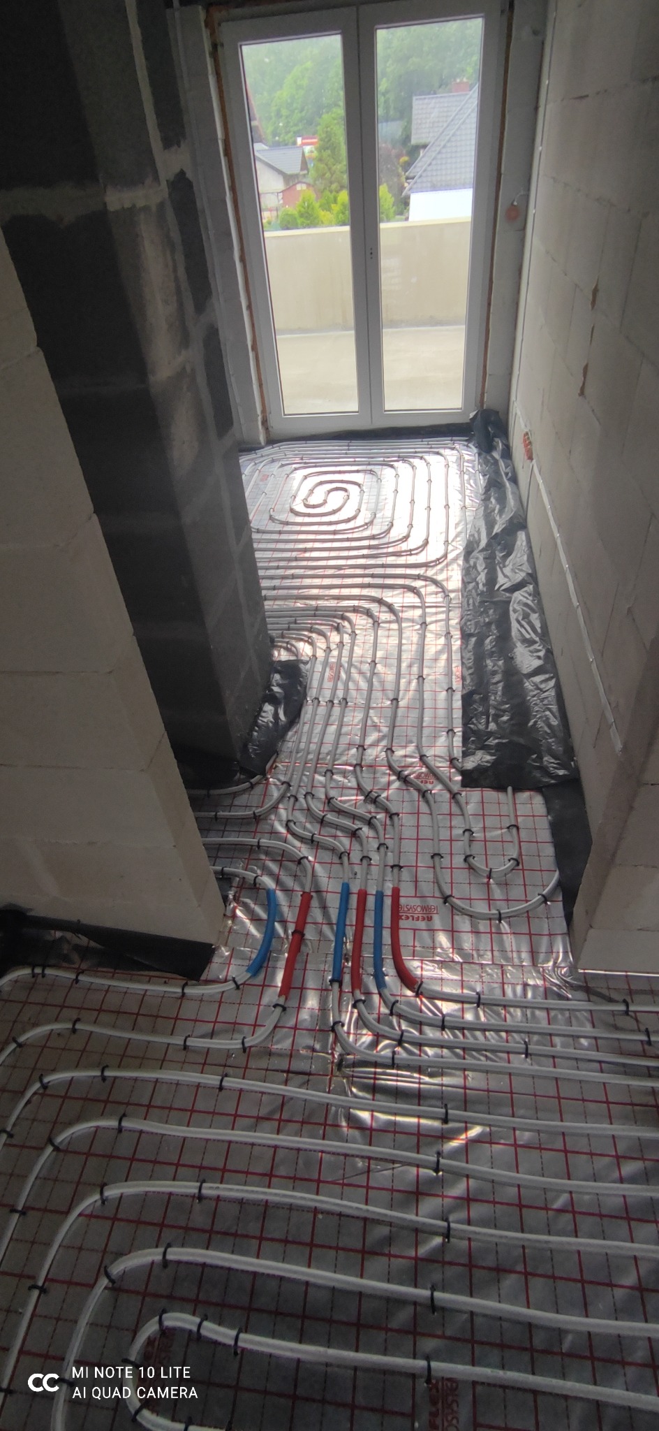 Montaż ogrzewania podłogowego w surowym stanie, widoczne rury grzewcze ułożone na folii z siatką, przyłącza rur w kolorze niebieskim i czerwonym, ściany w trakcie budowy, widok na zewnątrz przez...