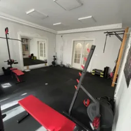 Domowa siłownia z czerwonymi ławkami, czarną podłogą i różnorodnym sprzętem do ćwiczeń, widoczne lustro odbijające dodatkowy sprzęt.