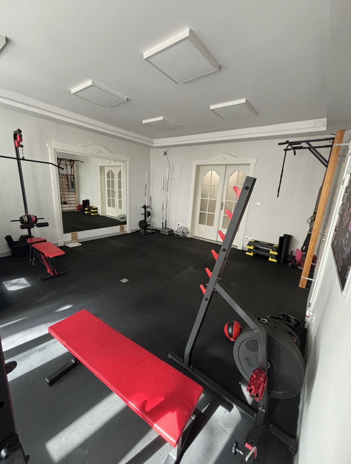 Domowa siłownia z czerwonymi ławkami, czarną podłogą i różnorodnym sprzętem do ćwiczeń, widoczne lustro odbijające dodatkowy sprzęt.