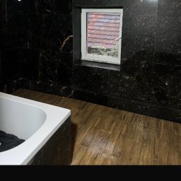 S.EnergyBud Dawid Suduł - Wykończona łazienka z wanną, okienkiem i ciemnymi płytkami imitującymi marmur na ścianach i podłodze. Widok z góry na dach budynku za oknem.