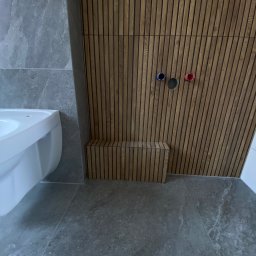 S.EnergyBud Dawid Suduł - Nowoczesna łazienka: szara płytka, drewniane lamele na ścianie z widocznymi przyłączami wodnymi i biała umywalka. Minimalistyczny design.