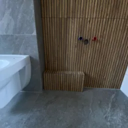 Nowoczesna łazienka: szara płytka, drewniane lamele na ścianie z widocznymi przyłączami wodnymi i biała umywalka. Minimalistyczny design.