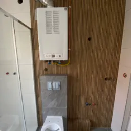 Nowoczesna łazienka z kabiną prysznicową, wiszącą toaletą i piecem gazowym na ścianie z drewnianymi lamelami. Widoczne miedziane rury i szara płytka podtynkowa.