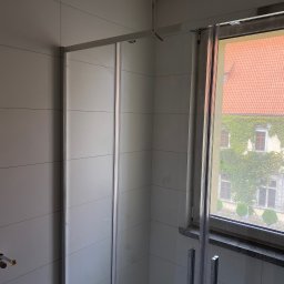 S.EnergyBud Dawid Suduł - Wykończona łazienka z kabiną prysznicową, jasnymi płytkami i oknem z widokiem na budynek porośnięty bluszczem. Widoczne niedokończone podłączenie kabla.