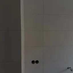 Ściana wyłożona jasnymi, kwadratowymi płytkami z widocznymi otworami na instalacje i fragmentem rurki, ukazująca etap prac wykończeniowych w łazience.