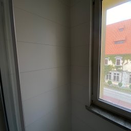 S.EnergyBud Dawid Suduł - Wykończona ściana w łazience z widokiem na budynek z ceglanym dachem przez okno. Białe panele ścienne z delikatnymi liniami. Granitowy parapet.