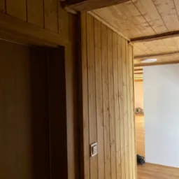 Wykończone wnętrze z drewnianą boazerią ścienną i sufitową, widoczny fragment drzwi i podłogi. Nowoczesny włącznik światła na ścianie. Estetyczne wykończenie w drewnie.