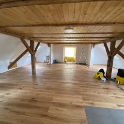 S.EnergyBud Dawid Suduł - Jasne panele podłogowe w przestronnym, drewnianym poddaszu z widocznymi belkami stropowymi i elementami wyposażenia, takimi jak drabina i krzesło.
