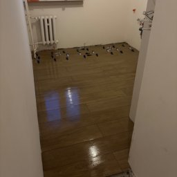 S.EnergyBud Dawid Suduł - Układanie brązowych płytek imitujących drewno w łazience, widoczne niebieskie krzyżyki dystansowe, grzejnik i okno w tle. Częściowo wykończona podłoga.