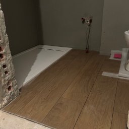 S.EnergyBud Dawid Suduł - Układanie płytek imitujących drewno w łazience w Lwówku Śląskim. Widoczny brodzik i fragment toalety. Prace w toku, surowe ściany i instalacje.